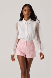A woman is wearing a white button-up shirt with sheer, long, puffed sleeves made of a transparent fabric, and white cuffs. She pairs it with high-waisted pink plaid shorts.